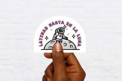 Hand holding a sticker with an astronaut graphic and "latinas hasta en la luna" text on a white background