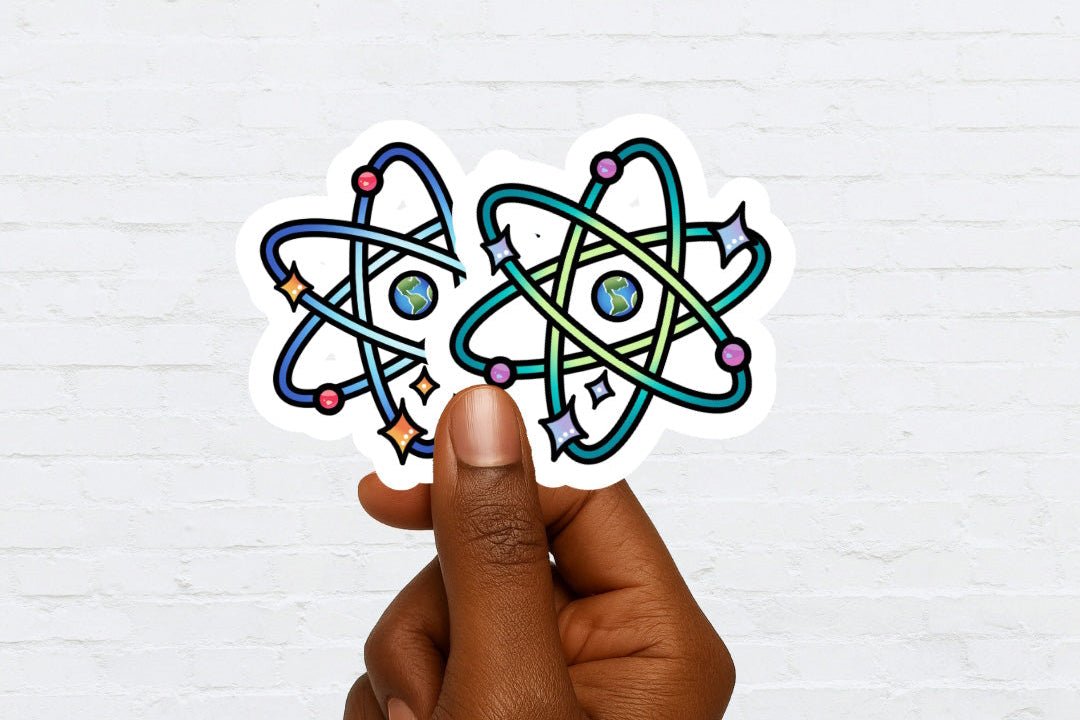 Hand holding 2 stickers with atom designs against a white brick wall background